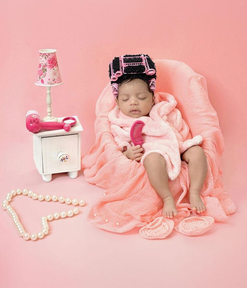 Sleeping newborn baby girl in a pink spa day setup, wrapped in a soft pink blanket and wearing multiple pink hair rollers, holding a pink comb while sitting in a fluffy pink chair with a pink lamp, pearl necklace, and pink slippers nearby.