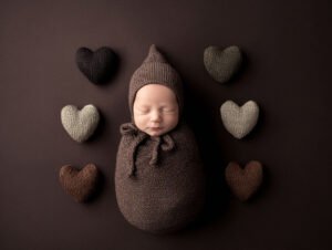 Peaceful newborn baby sleeping wrapped in a soft brown knitted cocoon surrounded by handmade knitted hearts in a warm studio setting