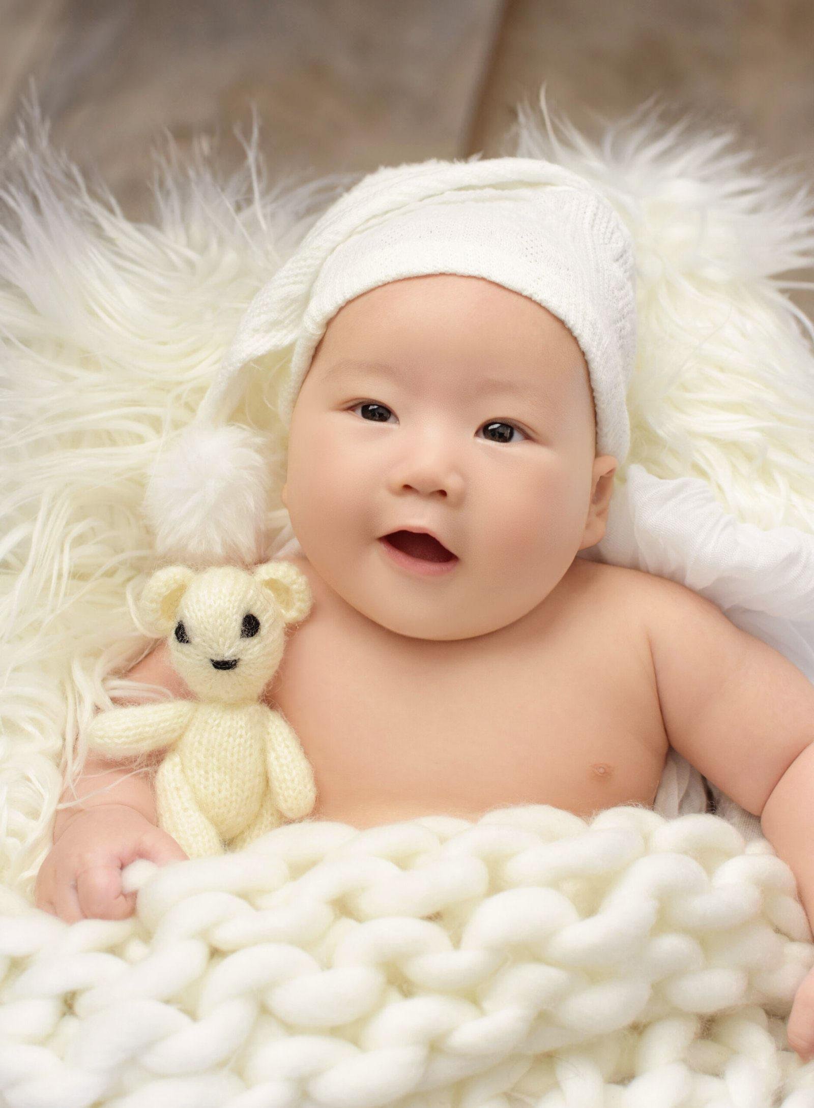 Awake newborn baby wearing a white knit bonnet, lying on a fluffy white rug and holding a small cream knitted teddy bear while looking directly at the camera with an open mouth.