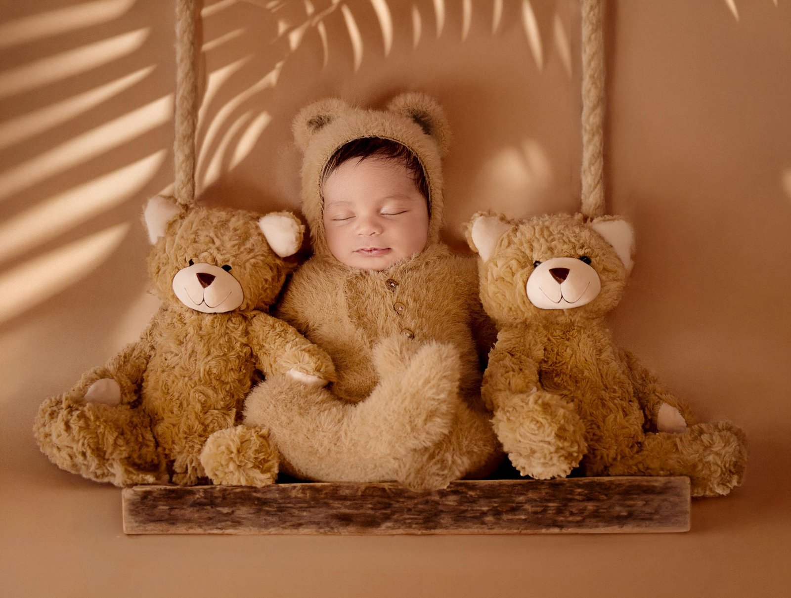 Sleeping newborn baby in a fluffy brown teddy bear onesie sitting on a wooden swing between two large brown teddy bears, with soft palm leaf shadows on the warm neutral background.