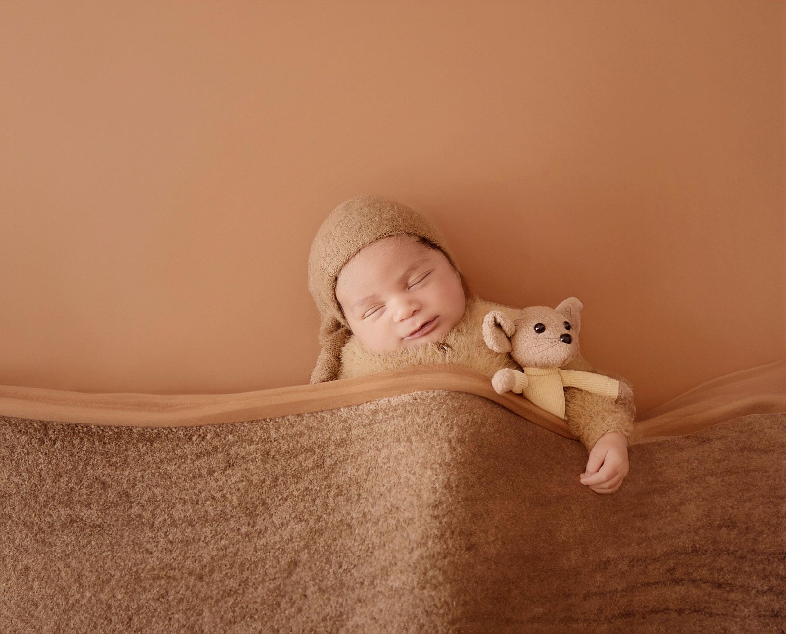 Sleeping newborn baby in a soft brown teddy bear onesie and matching bonnet, resting on a brown textured surface next to a small knitted teddy bear wearing a yellow sweater.