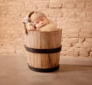 Sleeping newborn baby in a soft brown fuzzy outfit and bonnet, nestled inside a rustic wooden bucket with a small gray knitted mouse toy beside their head, against a light brick wall background.