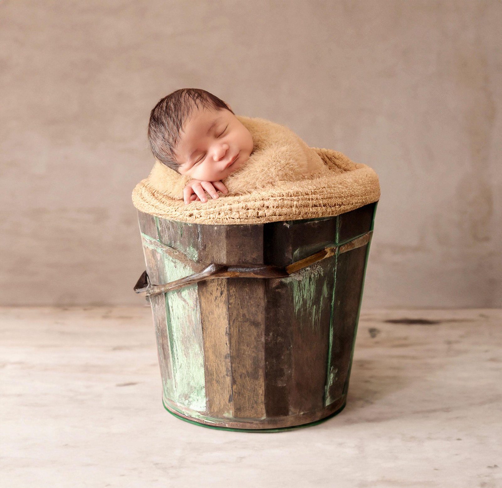 Sleeping newborn baby wrapped in a soft beige fuzzy blanket, resting their head on the rim of a rustic green and brown wooden bucket with one tiny hand visible.