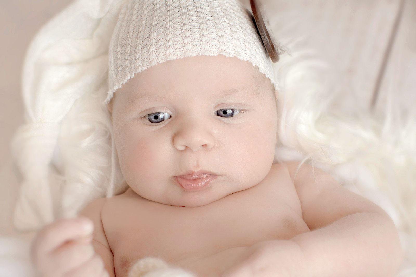 Close-up of an awake newborn baby wearing a white knit bonnet, lying on a fluffy white surface and looking directly at the camera with bright blue eyes and a sweet, slightly pursed expression.