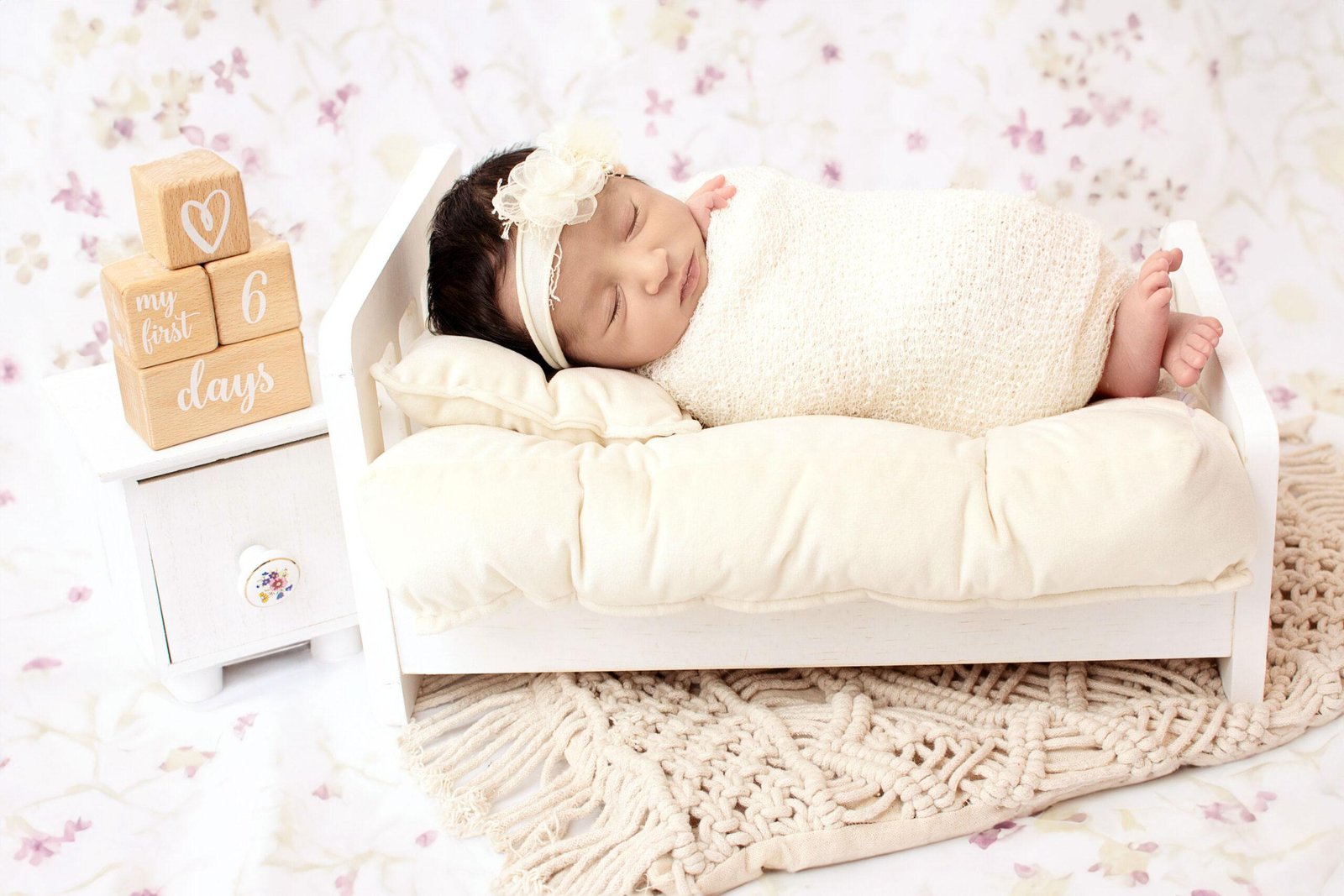 Sleeping newborn baby girl wrapped in a soft cream knit blanket, lying on her side in a miniature white wooden bed with a matching pillow, wearing a cream floral headband, next to wooden milestone blocks reading “my first 6 days” with a heart block on top.