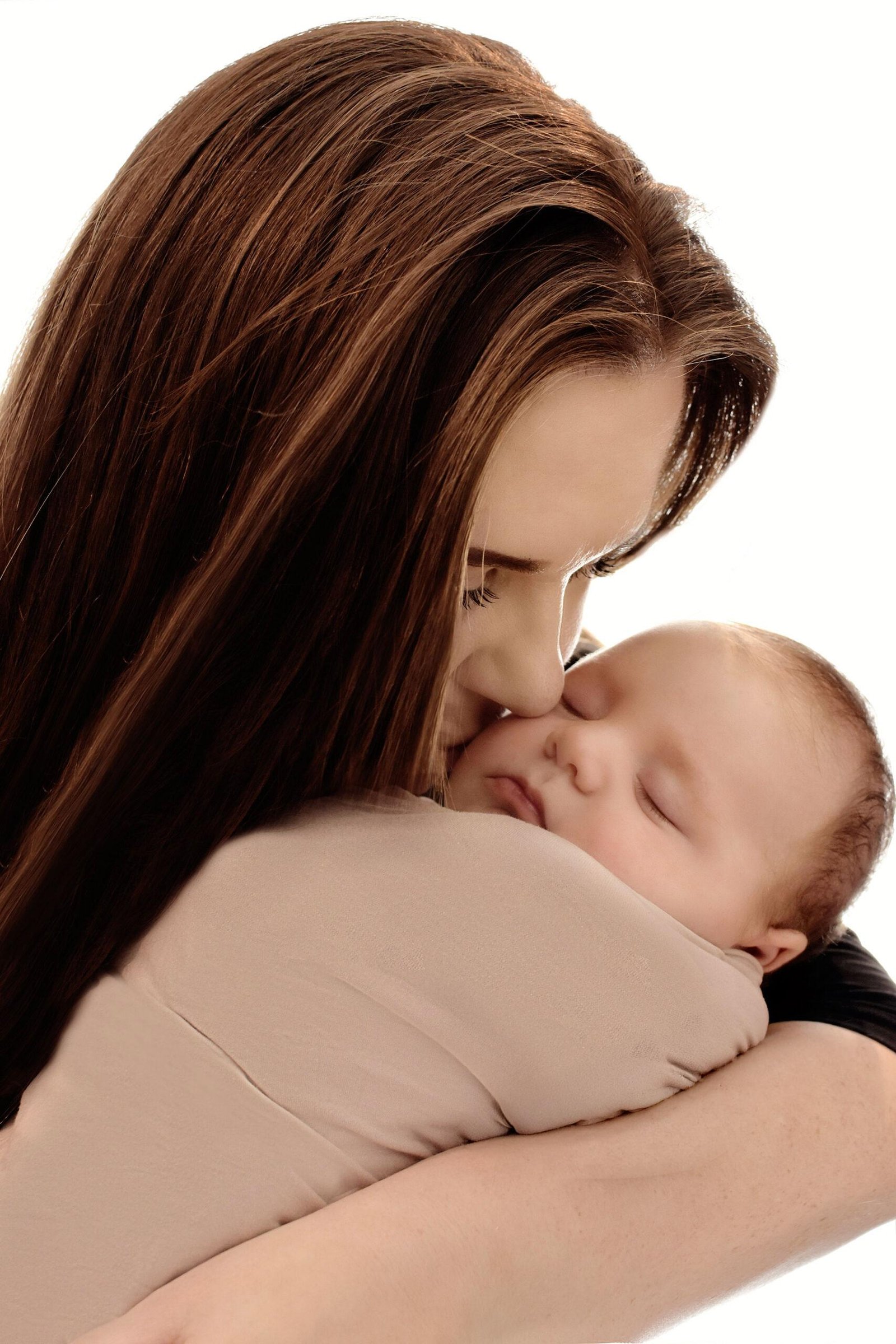 Tender moment of a mother with long brown hair gently kissing the forehead of her sleeping newborn baby, who is wrapped in a soft beige blanket and resting peacefully in her arms against a clean white background.