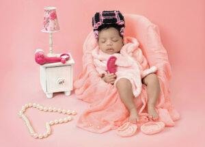 Sleeping newborn baby girl in a pink spa day setup, wrapped in a soft pink blanket and wearing multiple pink hair rollers, holding a pink comb while sitting in a fluffy pink chair with a pink lamp, pearl necklace, and pink slippers nearby.