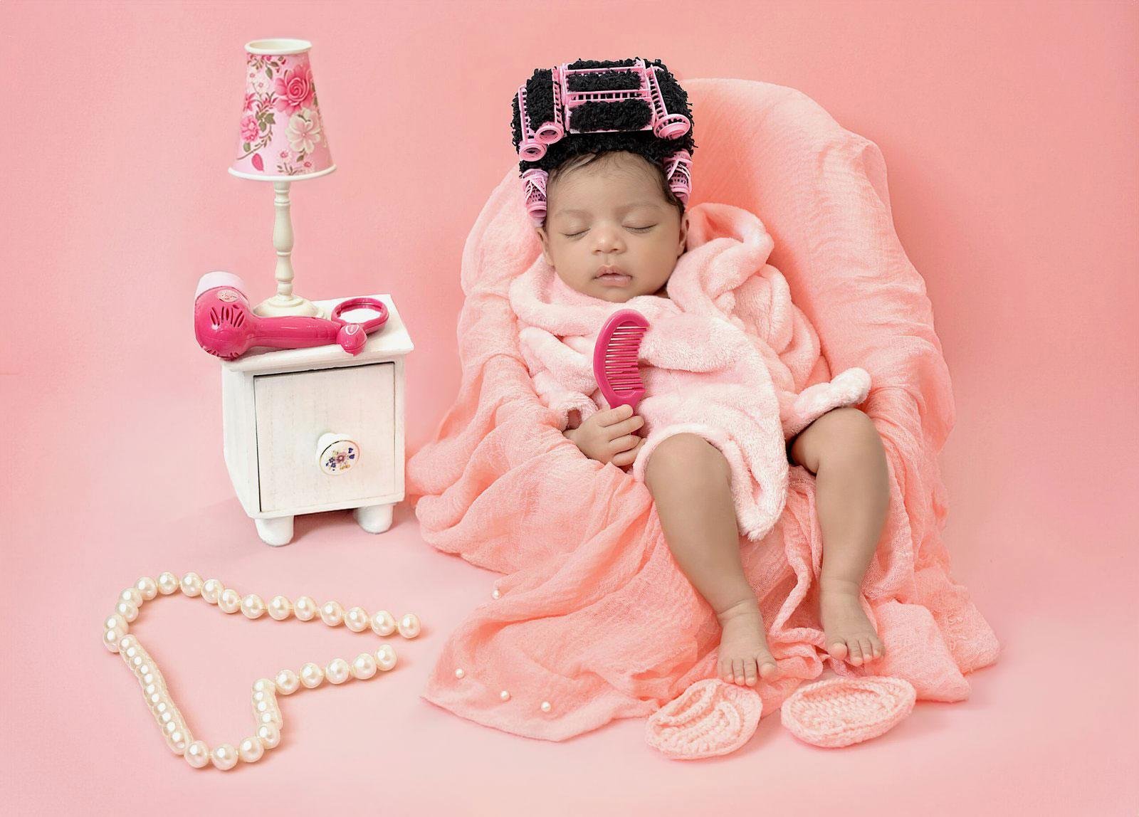 Sleeping newborn baby girl in a pink spa day setup, wrapped in a soft pink blanket and wearing multiple pink hair rollers, holding a pink comb while sitting in a fluffy pink chair with a pink lamp, pearl necklace, and pink slippers nearby.