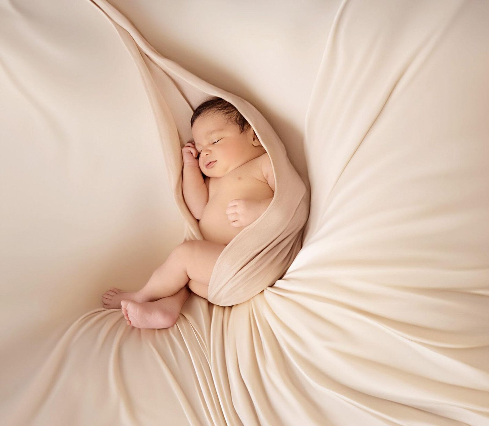 Sleeping newborn baby wrapped in soft beige fabric, nestled inside elegant flowing beige drapes arranged in a beautiful shape with one hand near the face and tiny feet visible.