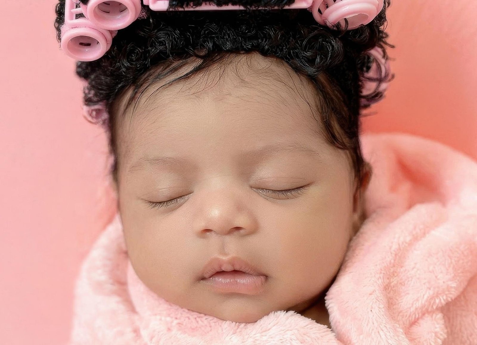 Close-up of a newborn baby girl with pink hair rollers and wrapped in a soft pink towel during spa day theme.