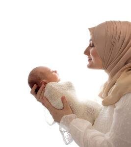 Mother wearing a beige hijab gently holding and looking lovingly at her newborn baby wrapped in a soft white knit blanket against a bright white background.