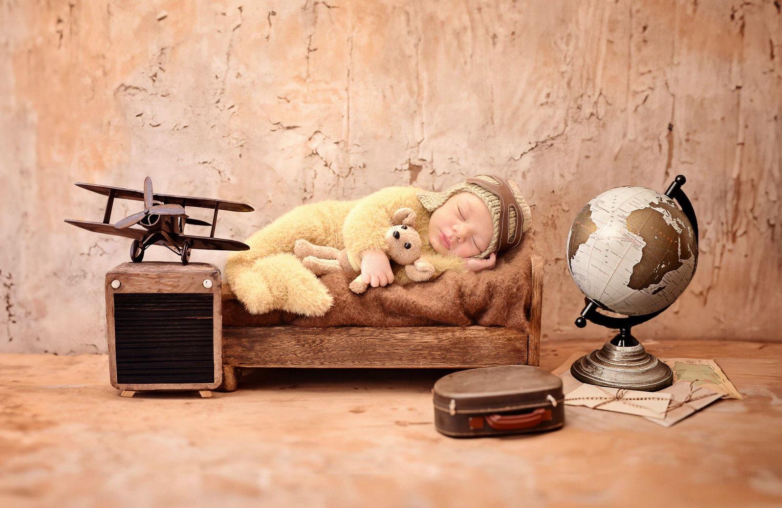 Sleeping newborn baby in a soft yellow fuzzy bear aviator onesie and knitted pilot hat with goggles, lying peacefully on a miniature wooden bed while cuddling a small teddy bear, surrounded by a vintage toy biplane, a globe, old suitcase, and travel maps.