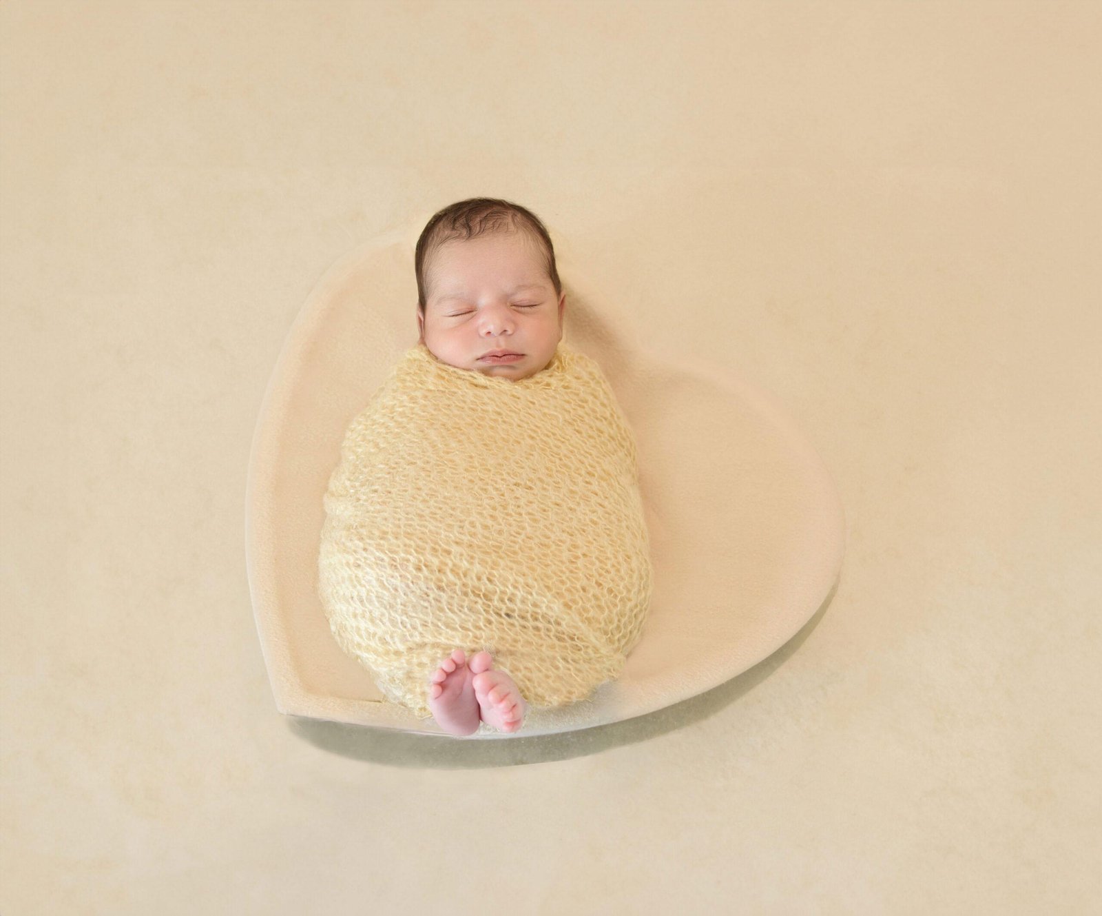 Sleeping newborn baby wrapped in a soft yellow knit blanket, lying peacefully inside a large white heart-shaped bowl with tiny feet peeking out at the bottom.