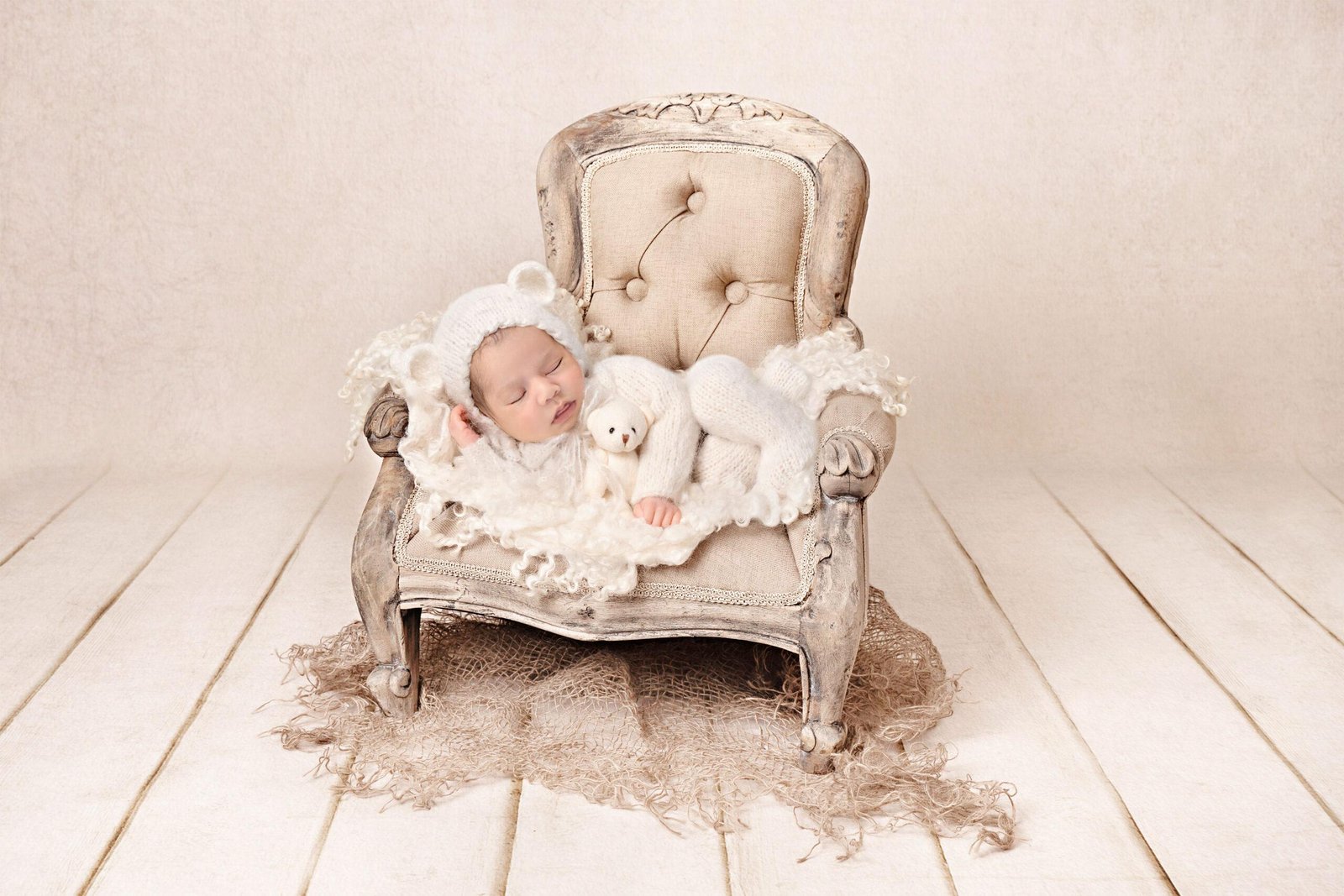 Sleeping newborn baby in a white fuzzy bear onesie with matching bonnet, lying peacefully on a vintage distressed white armchair while cuddling a small white teddy bear, surrounded by soft white tulle and burlap on a light wooden floor.