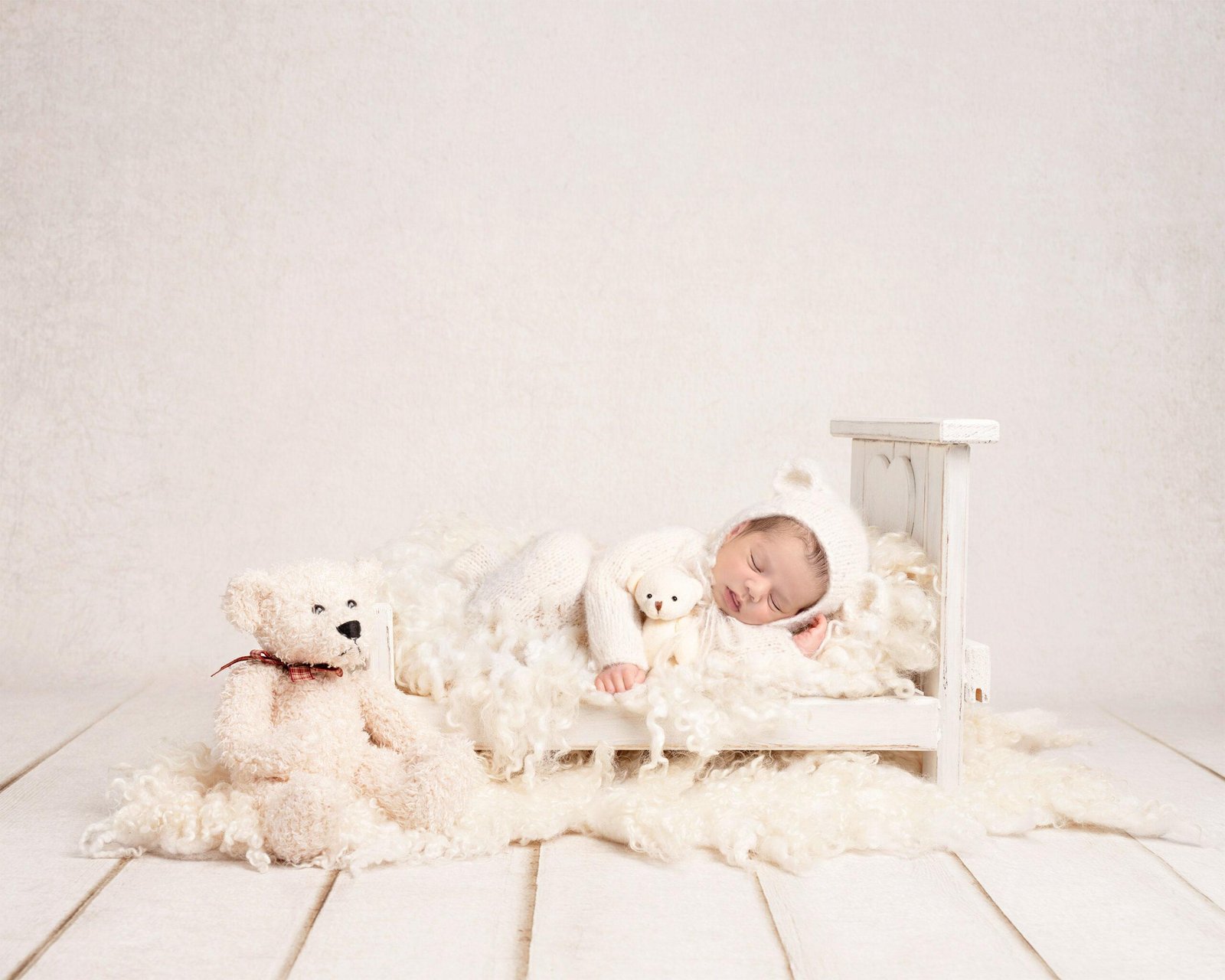 Sleeping newborn baby in a soft white knit outfit and matching bear-eared bonnet, lying peacefully on his side in a miniature white wooden bed while cuddling a small white teddy bear, with a large white teddy bear sitting nearby on a fluffy white rug.